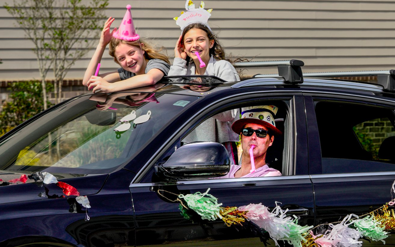 Man and two young girls wish friend a happy birthday with streamers and decorations while driving by in a car