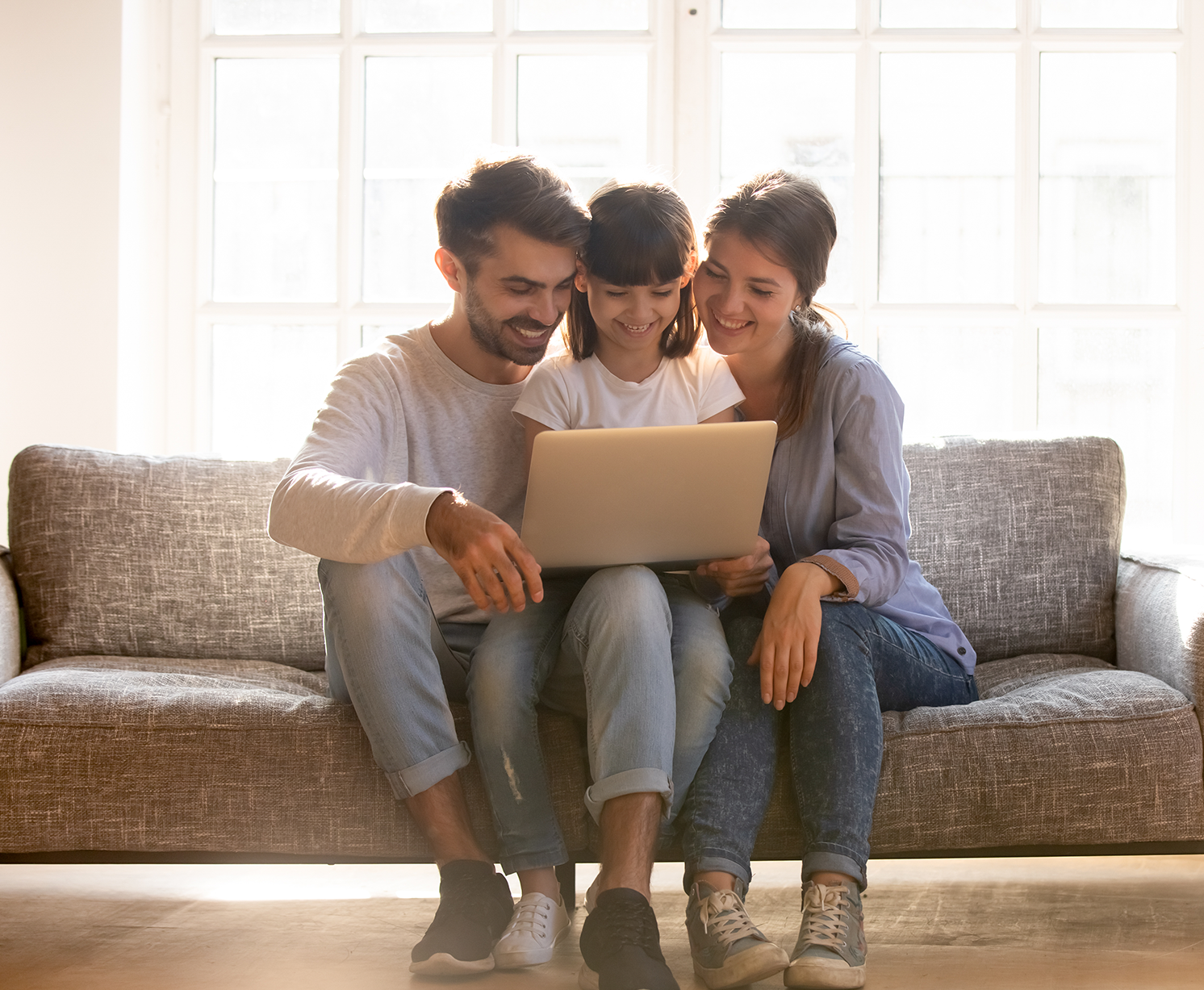 A mother, father, and daughter sit on the couch and smile while watching a video call on a laptop screen.